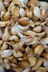 A close-up view of numerous roasted garlic cloves, some with papery skin partially peeled, piled together. The cloves vary in size and show a mix of light tan and golden brown hues, creating a textured, organic arrangement.