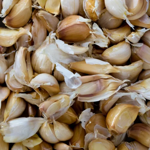 A close-up view of numerous roasted garlic cloves, some with papery skin partially peeled, piled together. The cloves vary in size and show a mix of light tan and golden brown hues, creating a textured, organic arrangement.