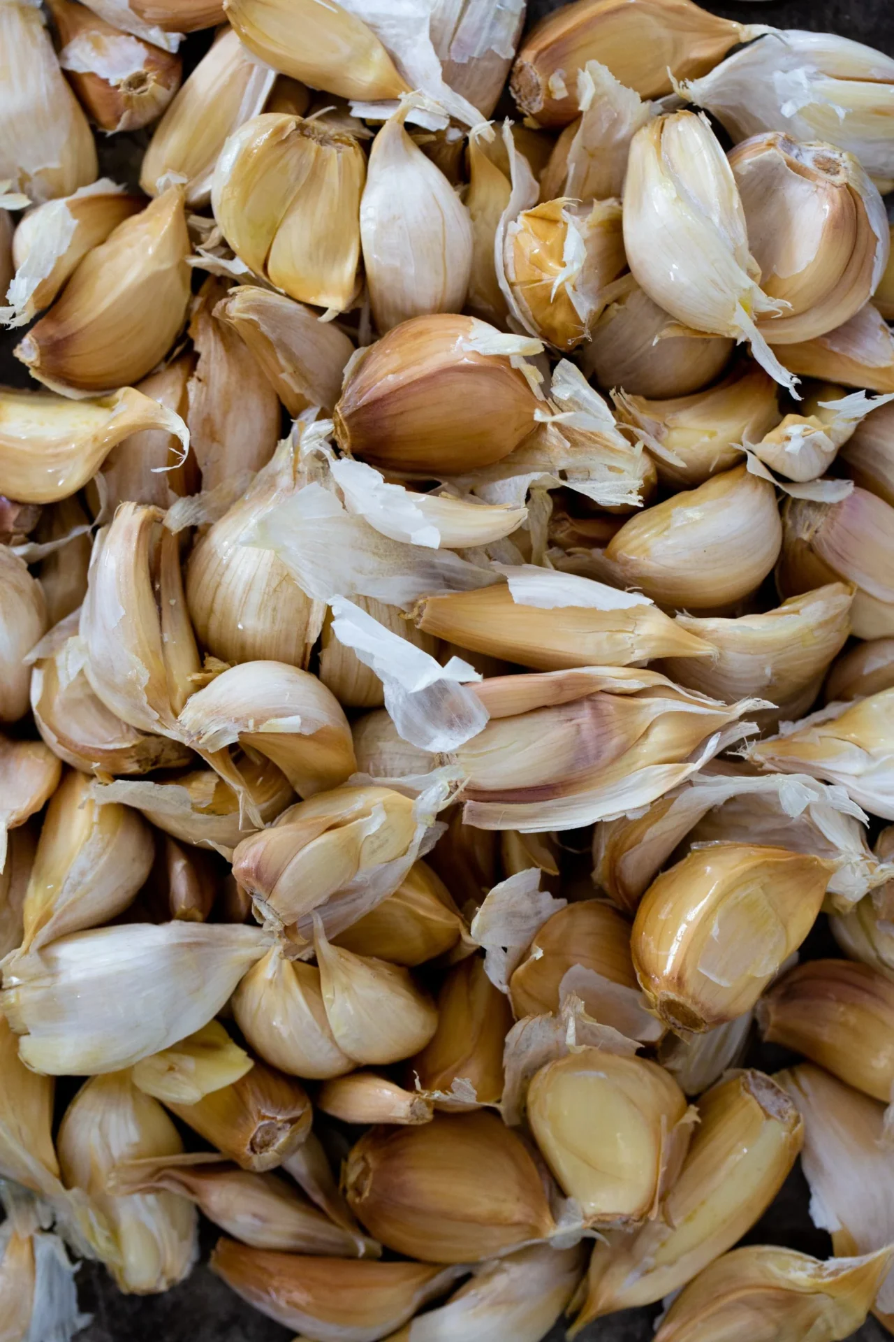 A close-up view of numerous roasted garlic cloves, some with papery skin partially peeled, piled together. The cloves vary in size and show a mix of light tan and golden brown hues, creating a textured, organic arrangement.