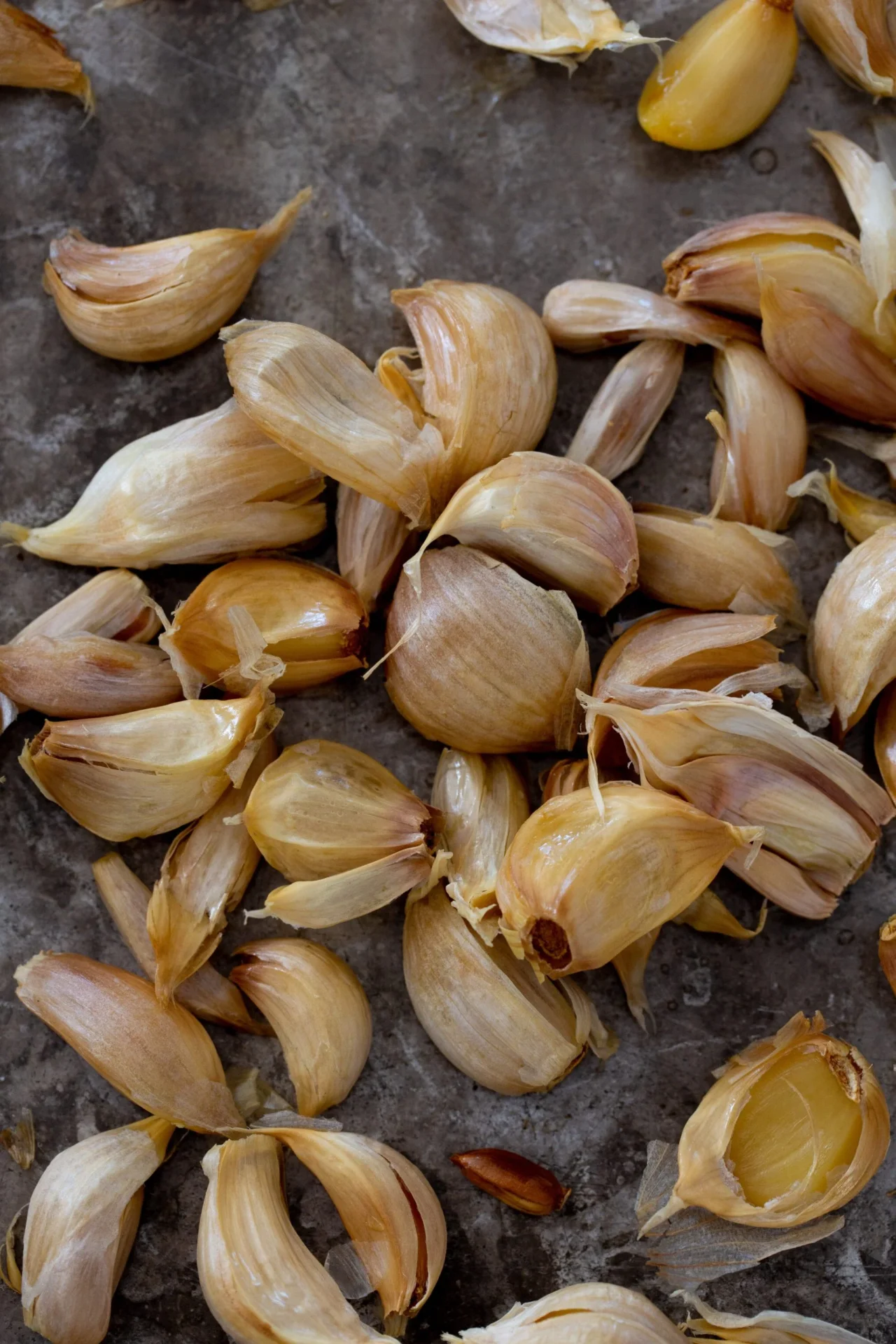 A close-up view of several roasted garlic cloves with papery skins partially peeled back, scattered on a rustic brown surface. The roasted garlic cloves are golden-brown, with caramelized edges and a soft, glossy appearance.