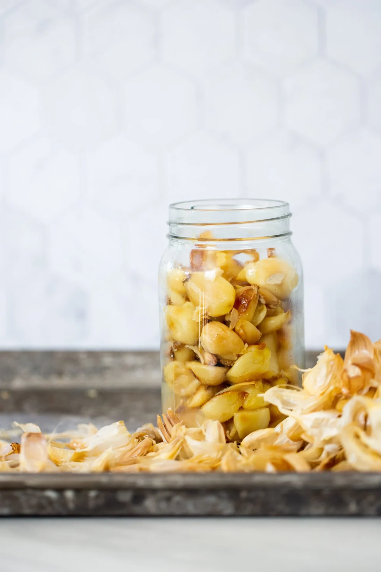 A glass jar filled with roasted garlic cloves sits on a tray. Scattered papery garlic skins lie in front of the jar. The light, hexagonal tile wall in the background adds to the clean and simple kitchen vibe, highlighting the golden roasted garlic.