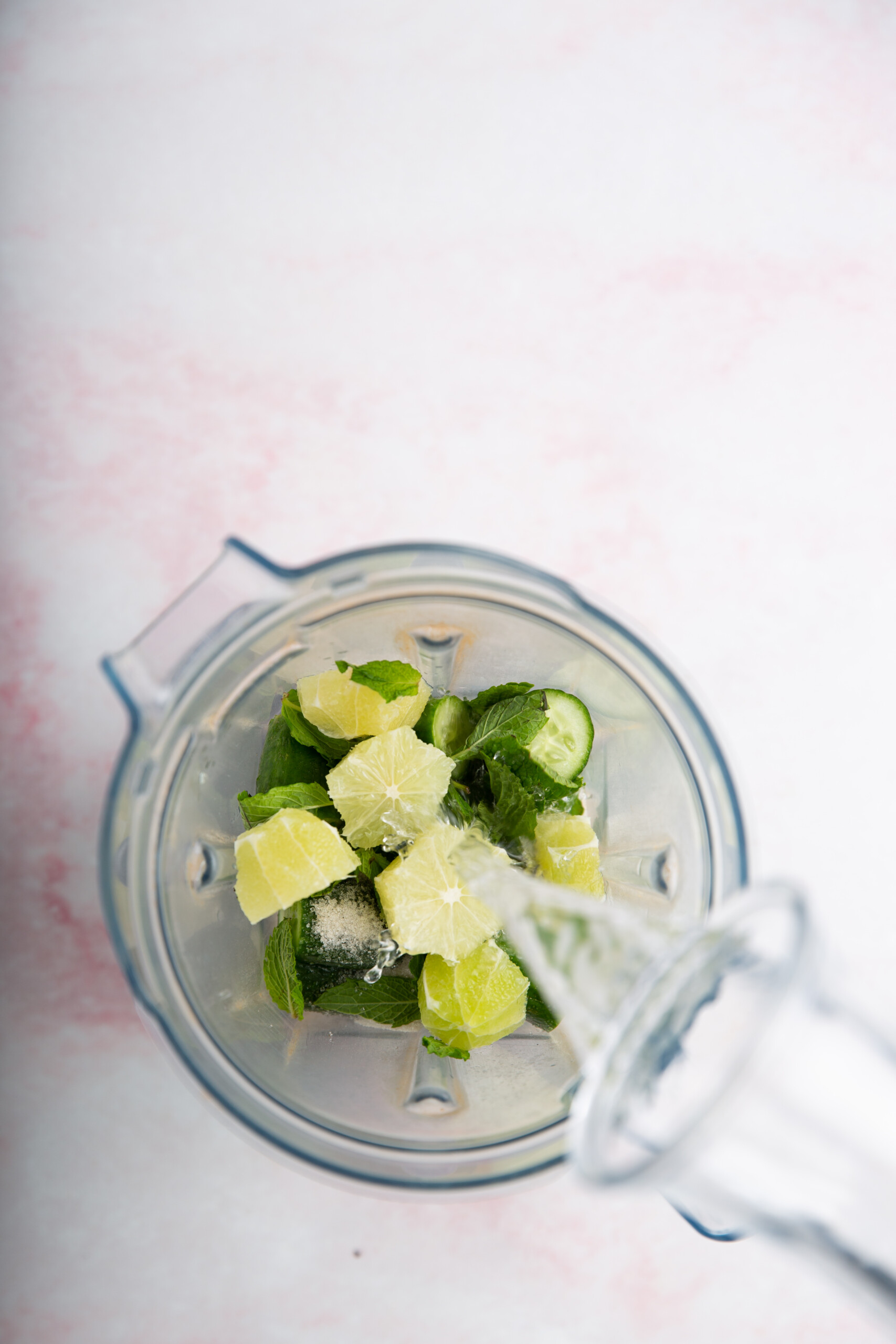 A top-down view of a blender containing lime wedges, cucumber slices, and mint leaves—perfect for pairing with totopos or sopa de frijol. Water is being poured in from a clear glass bottle. The background is a light pink and white surface.