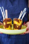 A person in a blue apron holds a yellow tray with two glass jars of swirled overnight oats topped with crumbly bits. Each jar features a wooden spoon and blue-striped paper straw. The person’s fingernails are painted red.