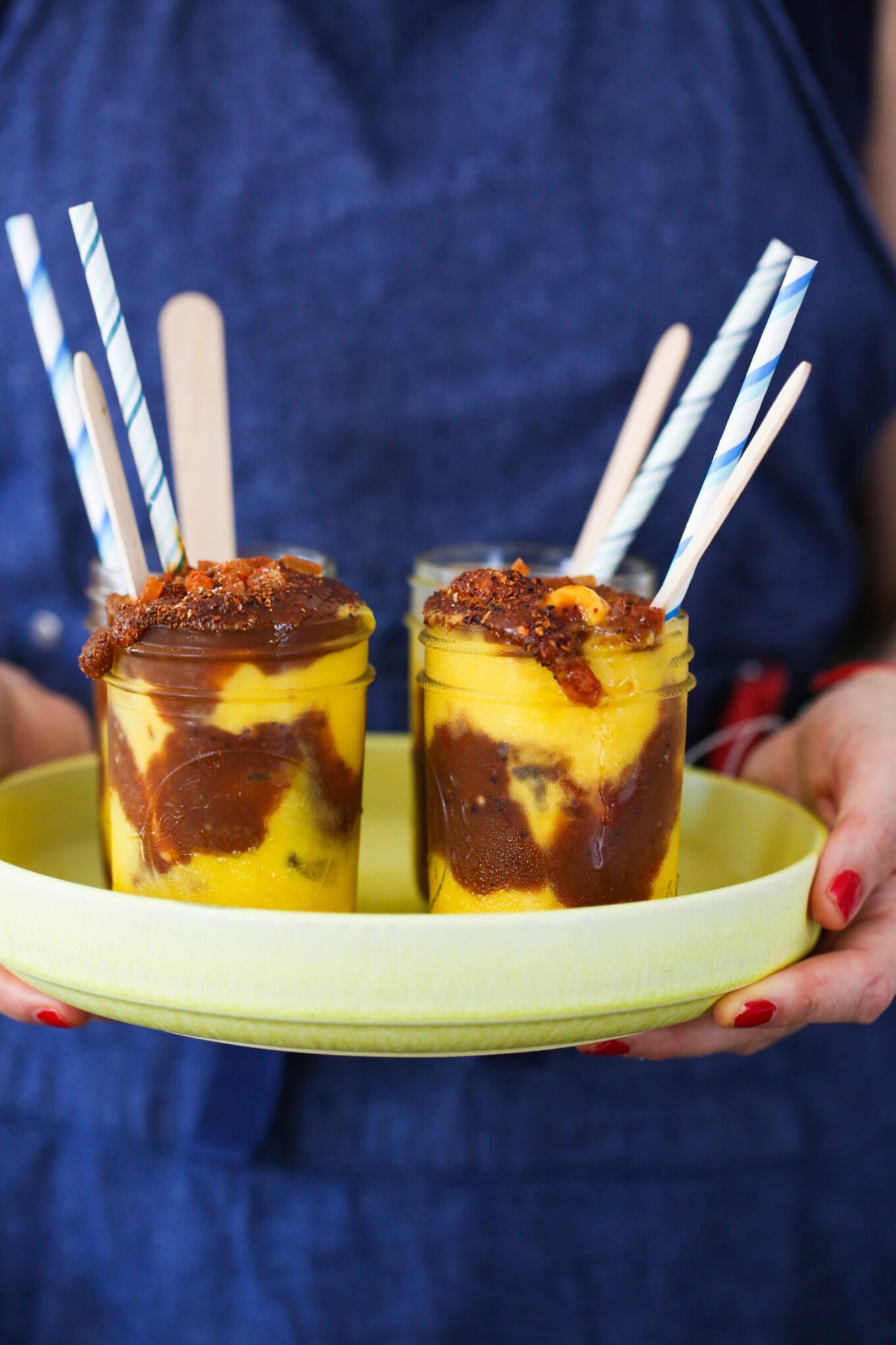 A person in a blue apron holds a yellow tray with two glass jars of swirled overnight oats topped with crumbly bits. Each jar features a wooden spoon and blue-striped paper straw. The person&rsquo;s fingernails are painted red.
