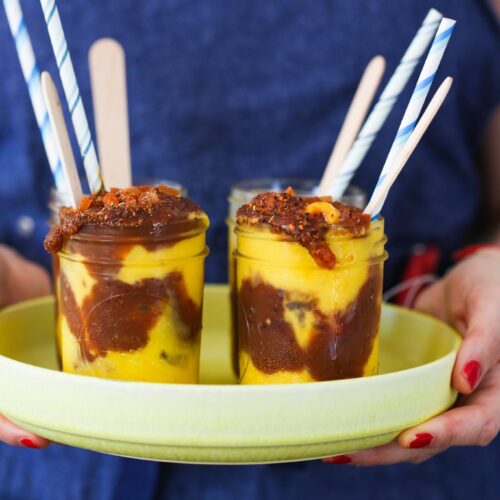 A person in a blue apron holds a yellow tray with two glass jars of swirled overnight oats topped with crumbly bits. Each jar features a wooden spoon and blue-striped paper straw. The person’s fingernails are painted red.