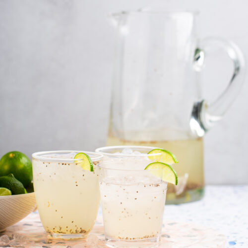 Two clear glasses filled with a light beige limonada con chia sit on a terrazzo tray, each garnished with a lime slice. A glass pitcher with more fresh drink is in the background, and a bowl of limes is on the side. The scene is set against a white textured background.