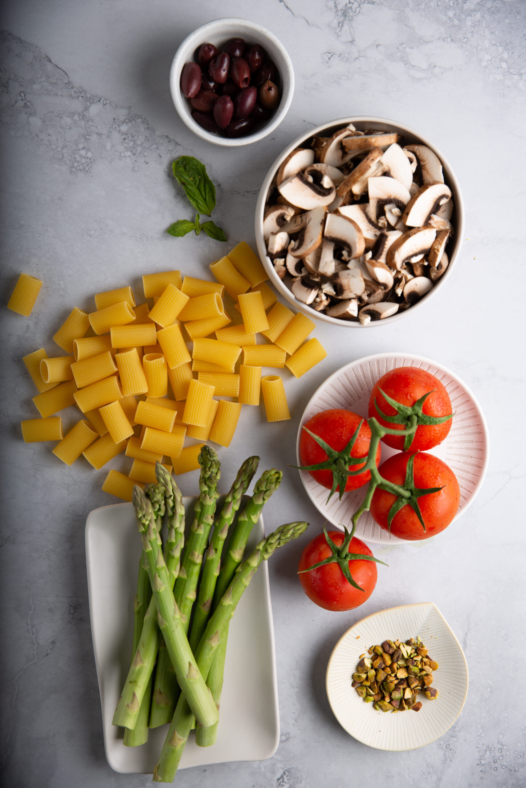 Overhead view of ingredients for pasta con esparragos on a gray surface: rigatoni, sliced mushrooms in a bowl, black olives in a small dish, whole tomatoes, fresh asparagus spears on a rectangular dish, pistachios, and a sprig of basil.
