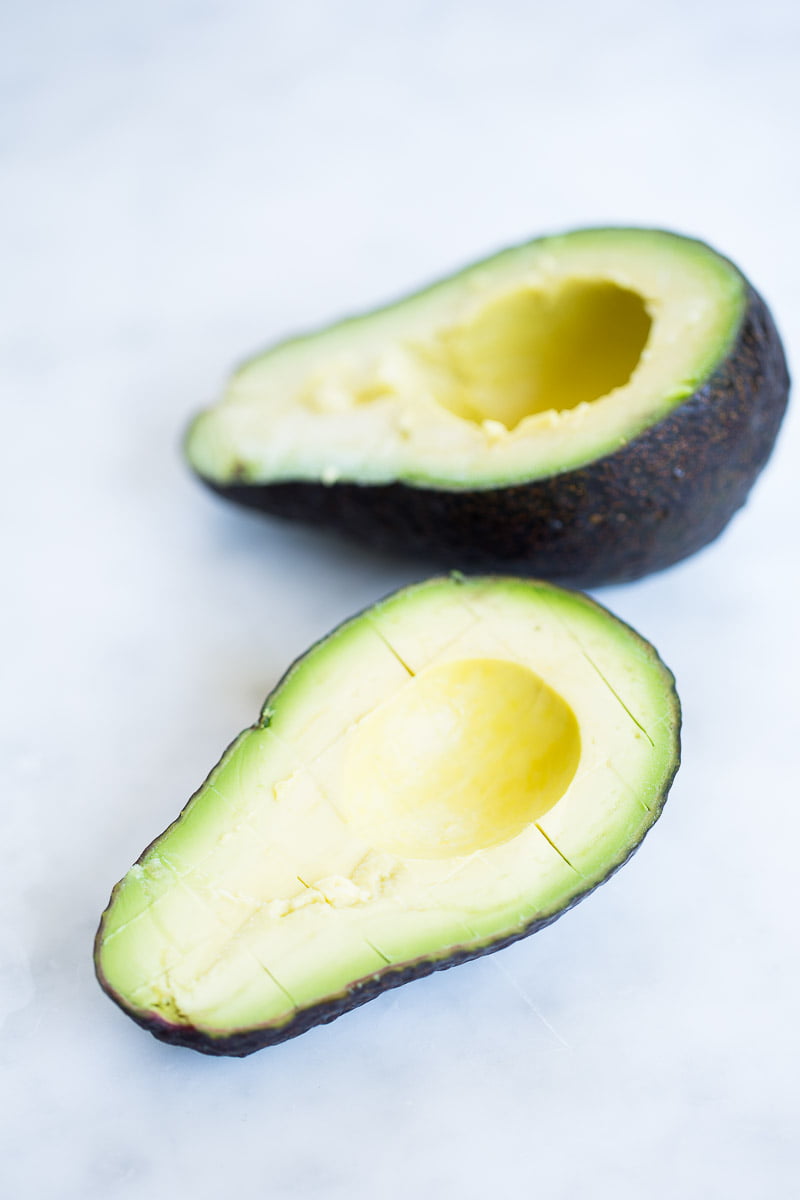 Two avocado halves rest on a white surface, the creamy foreground perfect for scooping into a quinoa summer salad. The blurred half behind also shows its pit removed and dark, textured skin.