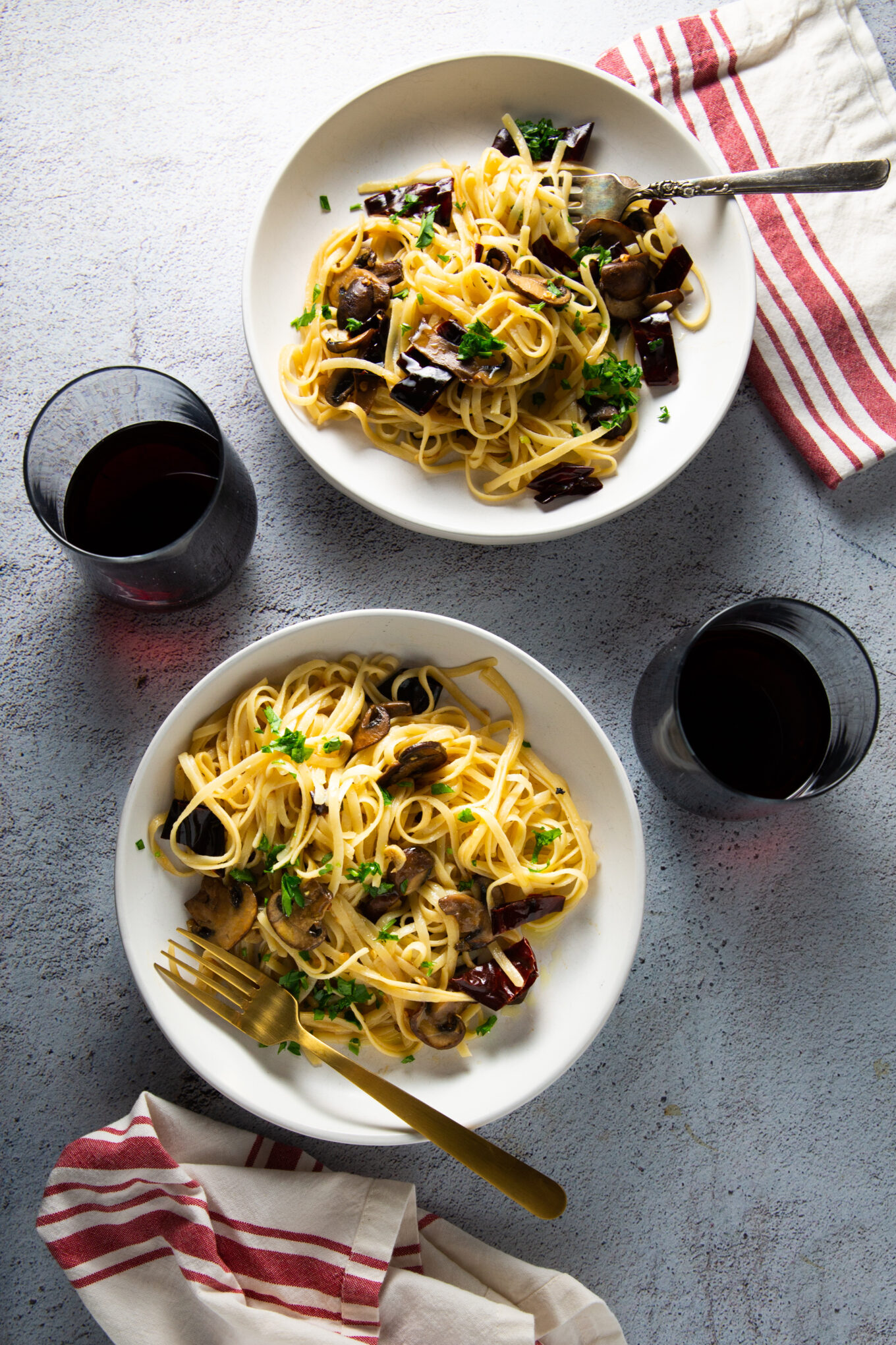 Two bowls of garlic mushroom pasta with sautéed vegetables, garnished with fresh herbs, sit on a light gray textured surface. Each bowl is accompanied by a fork and a napkin with red stripes. Two glasses of red wine are placed beside the bowls, adding to the elegant yet casual dining setup.