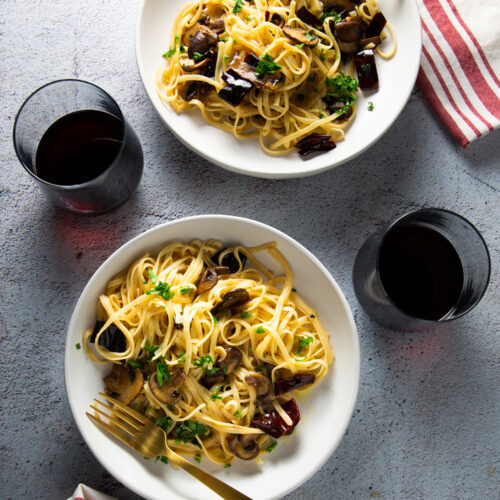 Two bowls of garlic mushroom pasta with sautéed vegetables, garnished with fresh herbs, sit on a light gray textured surface. Each bowl is accompanied by a fork and a napkin with red stripes. Two glasses of red wine are placed beside the bowls, adding to the elegant yet casual dining setup.
