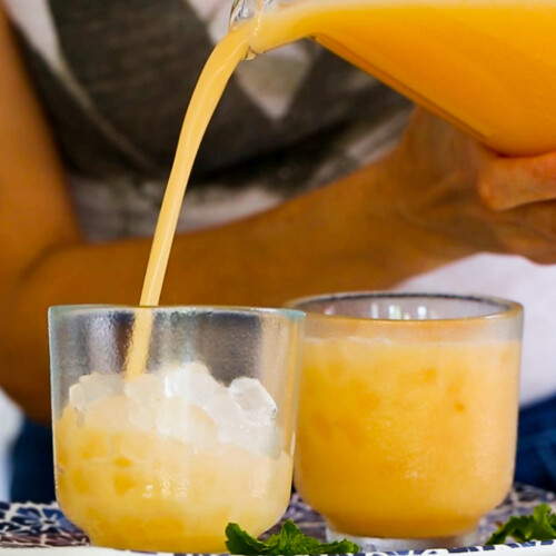 A person pours orange-colored agua de melon from a glass pitcher into a short glass filled with ice. Another filled glass sits beside it on a white tray with a blue pattern, garnished with fresh mint leaves. The background is softly blurred.