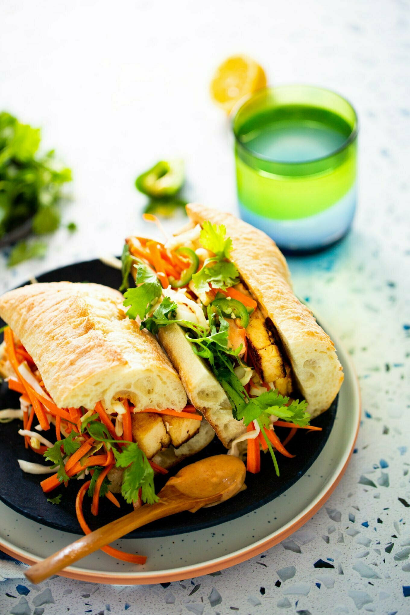 Un colorido sándwich con pollo a la parrilla, zanahorias ralladas, cilantro y jalapeños sobre rebanadas de baguette crujientes evoca la vibrante esencia de un bánh mì. Se coloca en un plato junto a una cuchara de madera llena de salsa. Cerca, un vaso con degradado verde y azul contiene agua, con hierbas y limón de fondo.