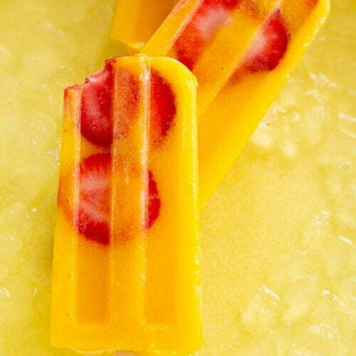 Three vibrant orange popsicles with embedded red strawberry slices and a hint of mango rest atop a bed of crushed ice. One popsicle has a bite taken out of it, revealing its creamy texture. The sunny yellow background enhances the bright, refreshing look of these fruity treats.
