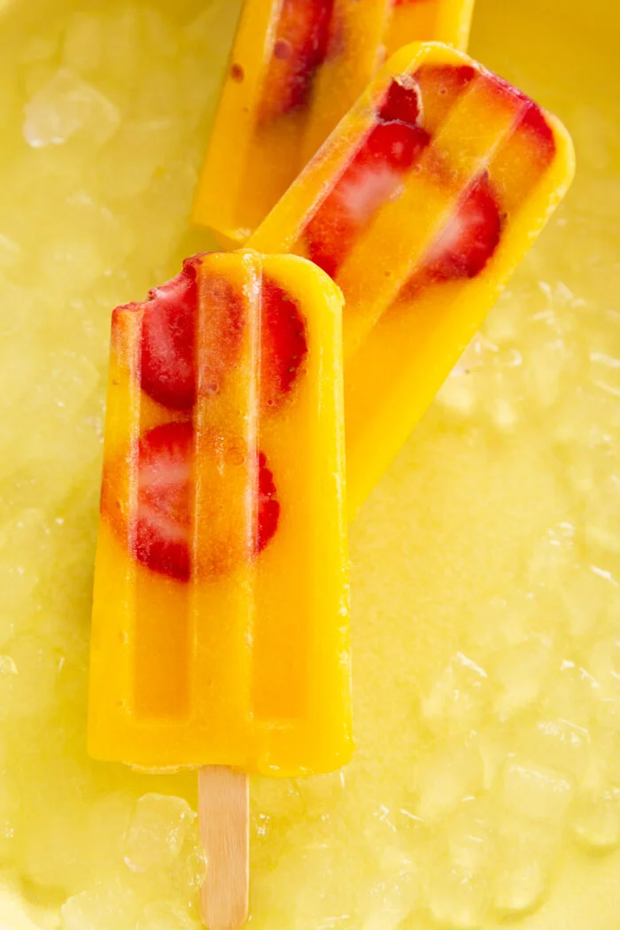 Three vibrant orange popsicles with embedded red strawberry slices and a hint of mango rest atop a bed of crushed ice. One popsicle has a bite taken out of it, revealing its creamy texture. The sunny yellow background enhances the bright, refreshing look of these fruity treats.
