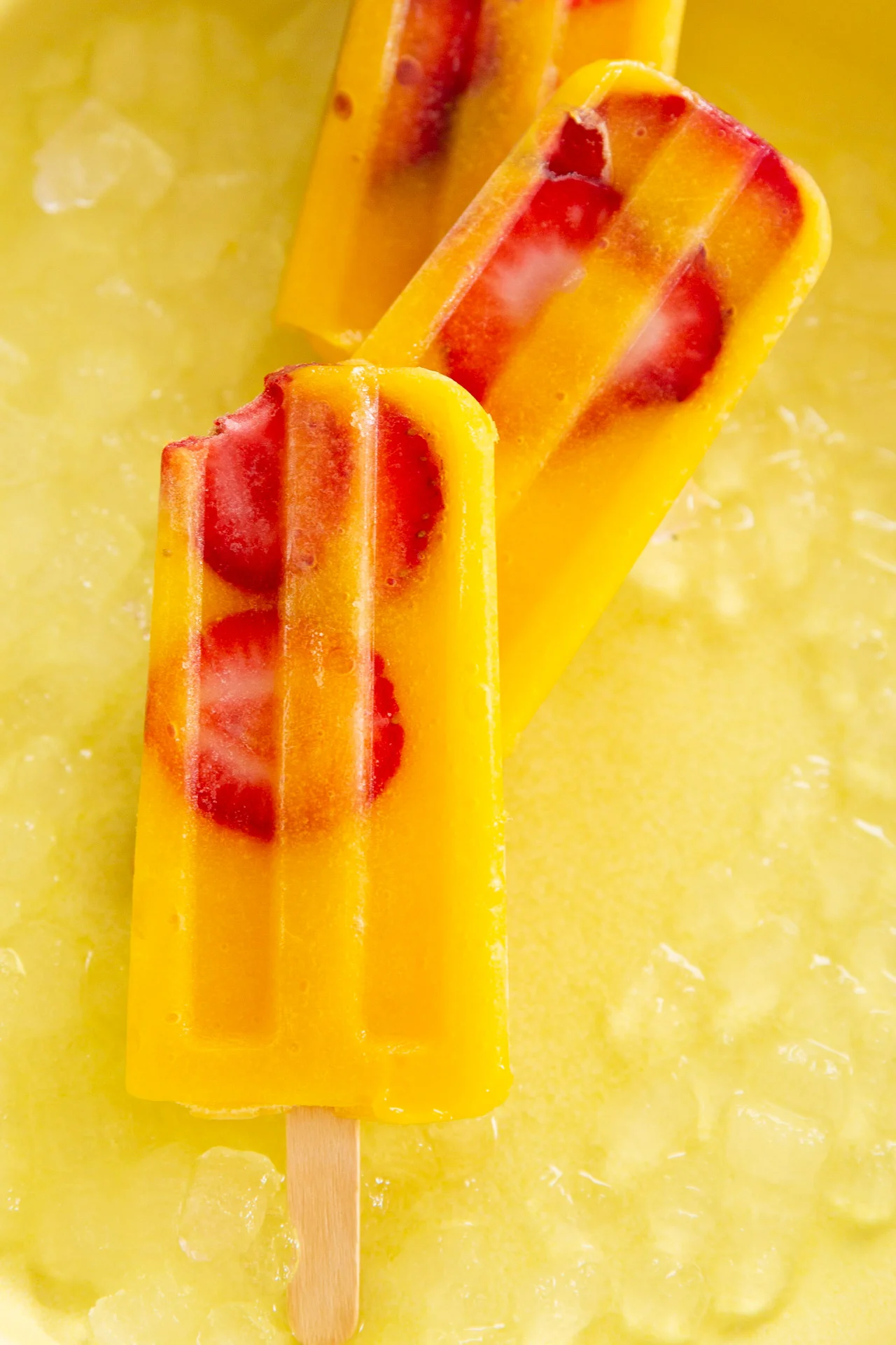 Three vibrant orange popsicles with embedded red strawberry slices and a hint of mango rest atop a bed of crushed ice. One popsicle has a bite taken out of it, revealing its creamy texture. The sunny yellow background enhances the bright, refreshing look of these fruity treats.