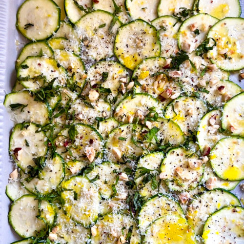 Thinly sliced zucchini arranged in overlapping layers on a white dish, topped with nuts, herbs, cheese, black pepper, and olive oil—this vibrant zucchini carpaccio is a perfect raw zucchini recipe for a fresh appetizer or salad.