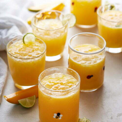 Six glasses of Mexican mango drink with ice are arranged on a white surface. Each glass is topped with lime slices and surrounded by lime wedges and orange slices. The drinks have a vibrant, refreshing appearance, and some glasses have tiny decorative prints on them.