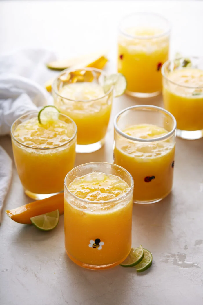 Six glasses of Mexican mango drink with ice are arranged on a white surface. Each glass is topped with lime slices and surrounded by lime wedges and orange slices. The drinks have a vibrant, refreshing appearance, and some glasses have tiny decorative prints on them.