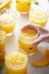 A hand is reaching for a glass of freshly squeezed orange juice, filled with ice and garnished with orange pulp. There are five other similar glasses of Mexican drink on a white surface, some with small fruit decorations. Mango slices and lime wedges are scattered around.