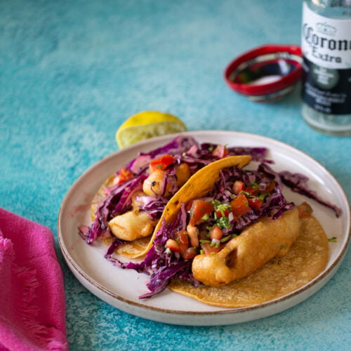 A beer-battered fish taco on a beige plate with a blue textured background. It’s loaded with red cabbage slaw and diced tomato salsa. A lime wedge sits beside it, while a pink napkin flanks the left and an open Corona beer garnishes the right, creating a perfect tacos night scene.