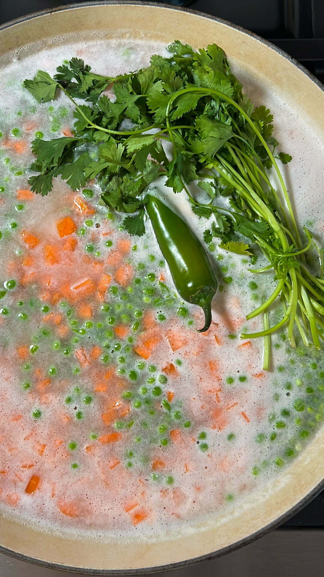 A pot of simmering soup with orange carrot cubes, green peas, foam on the surface, and fresh cilantro and a jalape&ntilde;o pepper resting on top, resembling a Mexican arroz Primavera in its early cooking stage with herbs and chili not yet mixed in.