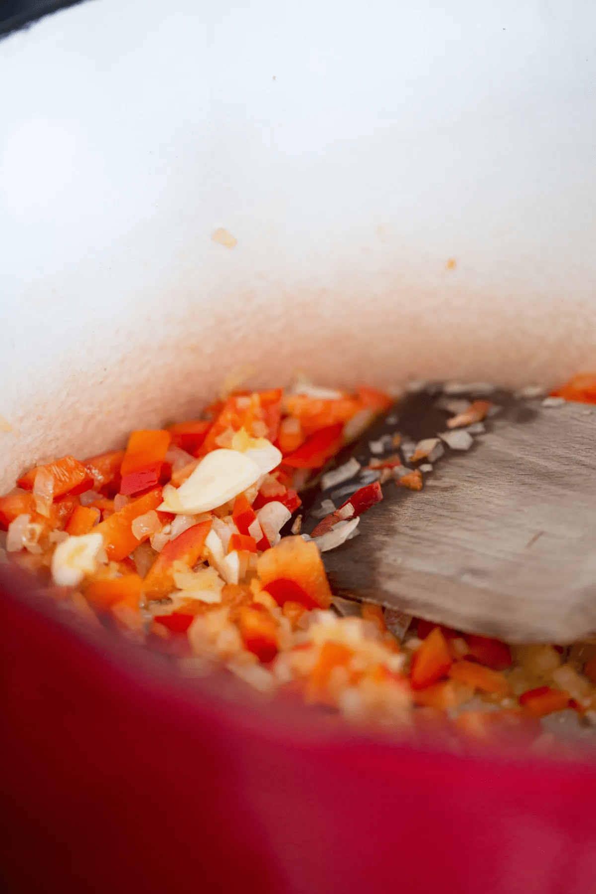 Diced red bell peppers, onions, and a clove of garlic are being sautéed in a red Dutch oven as the base for a hearty mushroom ragu. A wooden spatula stirs the colorful vegetables against the pot’s white interior.