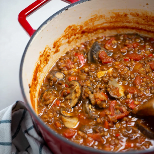 A red Dutch oven filled with a chunky lentil mushroom ragu featuring tomatoes and vegetables. The pot is stained with sauce and sits on a striped cloth, with a wooden spoon partially visible stirring the hearty mixture.