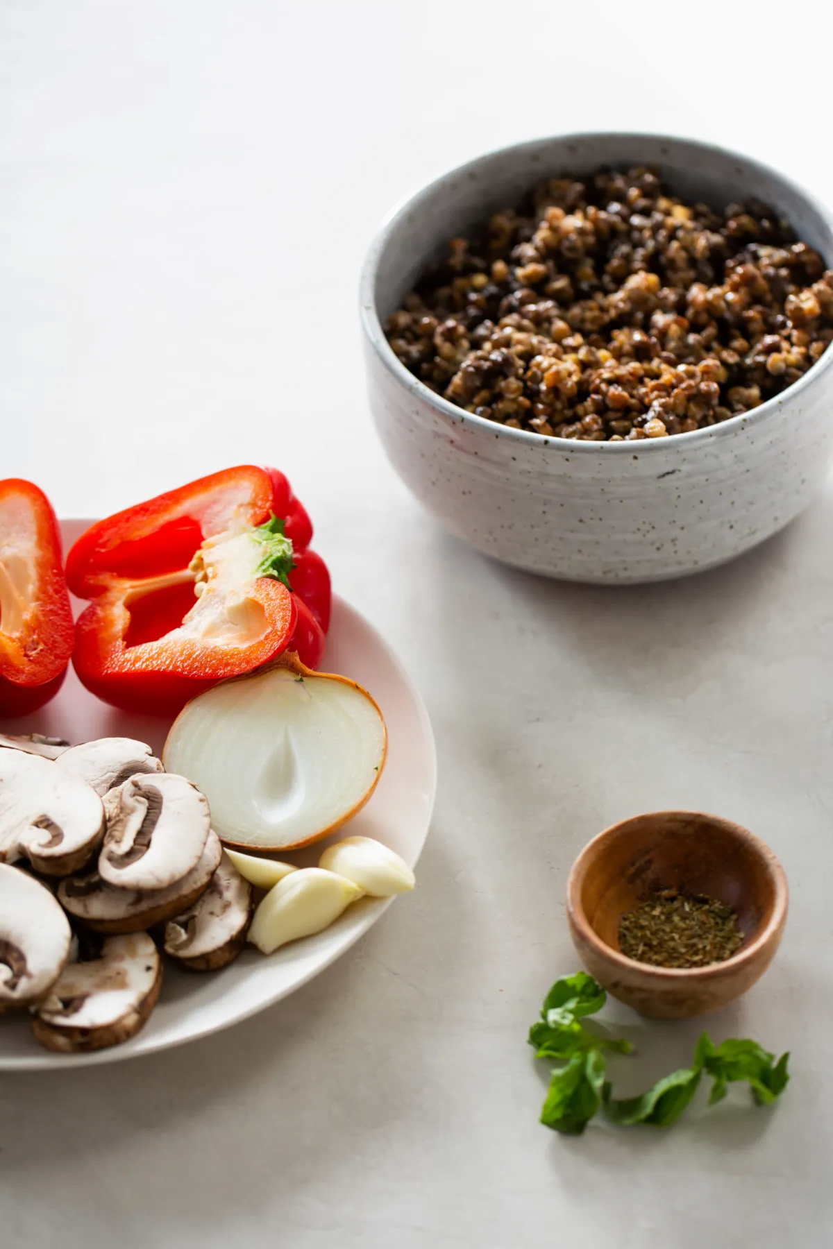 A white plate holds sliced mushrooms, half a red bell pepper, a halved onion, and two garlic cloves—perfect for a flavorful mushroom ragu. Nearby are cooked quinoa, dried herbs, and basil leaves on a light-colored surface.