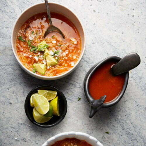 Two bowls of fideo soup garnished with avocado, cheese, and herbs. Nearby are lime wedges in a small black bowl and a dolphin-shaped sauce boat with additional soup. Each bowl has a spoon. The background is light gray, adding contrast to the vibrant tomato soup.