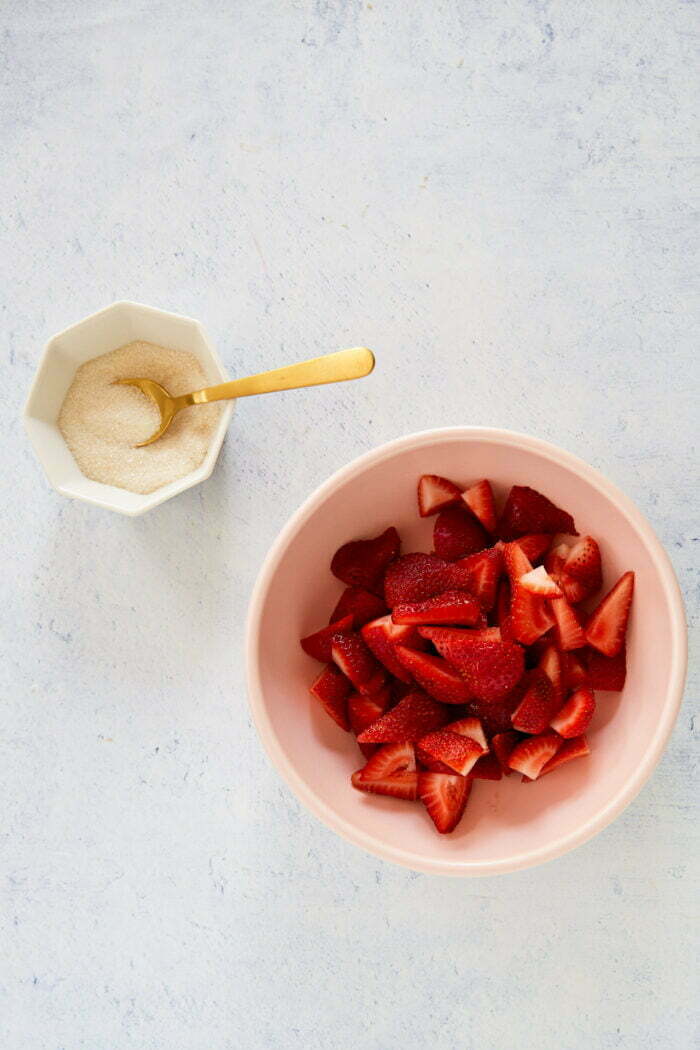 A bowl with sliced strawberries sits on a light surface, reminiscent of vegan fresas con crema. Next to it, a small hexagonal dish contains sugar with a gold spoon resting inside. The vibrant red strawberries pop in the bright and minimalist setting.
