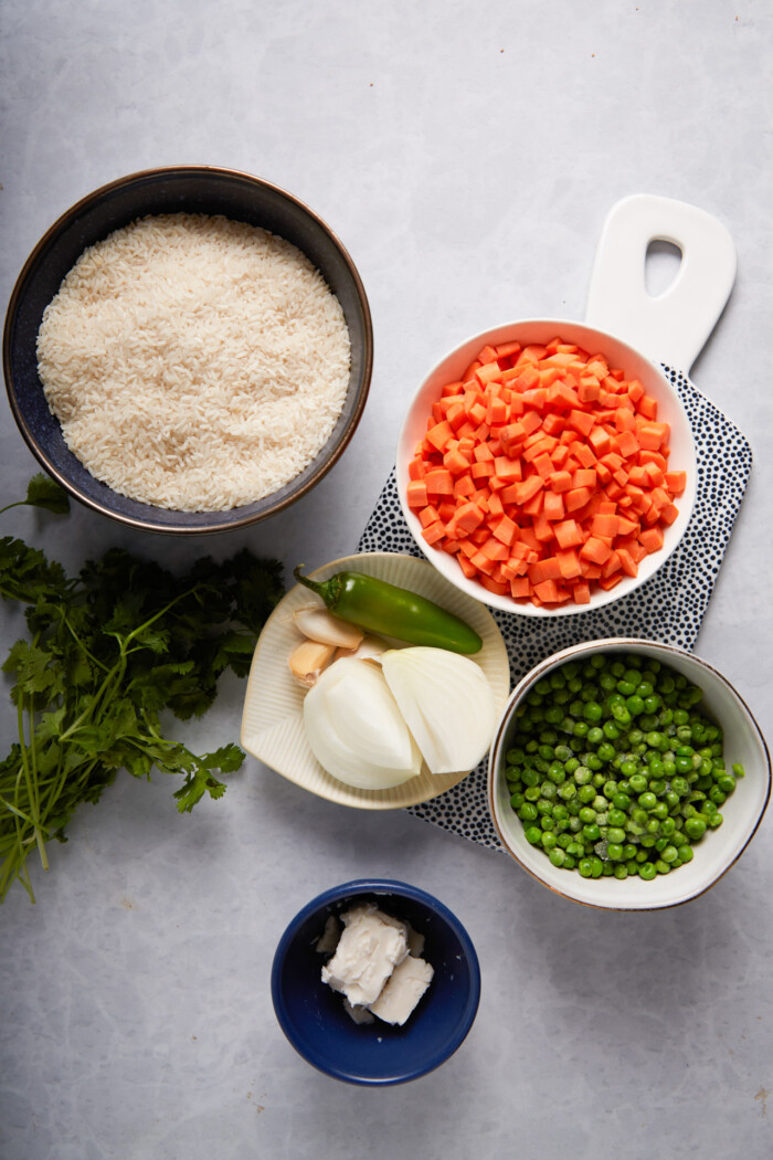 uncooked white rice in a bowl, chopped carrrots, frozen peas and onions and garlic cloves next to a bunch of cilantro.