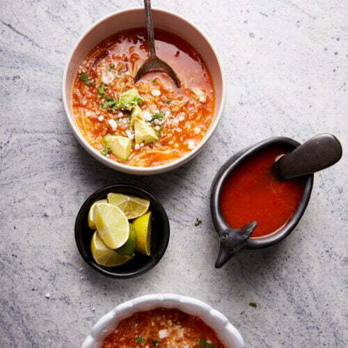 Top view of two bowls of tomato-based fideo soup garnished with avocado chunks and cheese, each with a spoon. Accompaniments include a small bowl with lime wedges and a sauce boat filled with red sauce with a black ladle. Everything is arranged on a light gray marble surface.