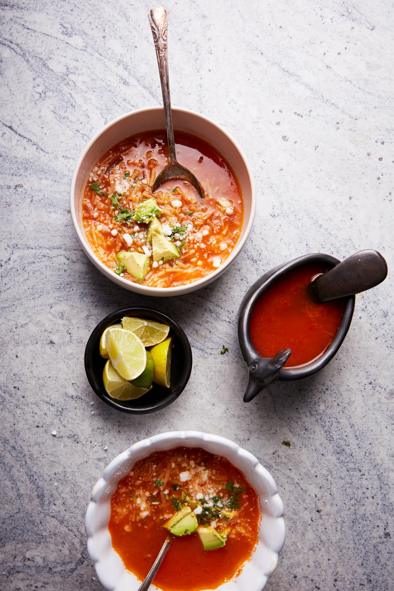 Top view of two bowls of tomato-based fideo soup garnished with avocado chunks and cheese, each with a spoon. Accompaniments include a small bowl with lime wedges and a sauce boat filled with red sauce with a black ladle. Everything is arranged on a light gray marble surface.