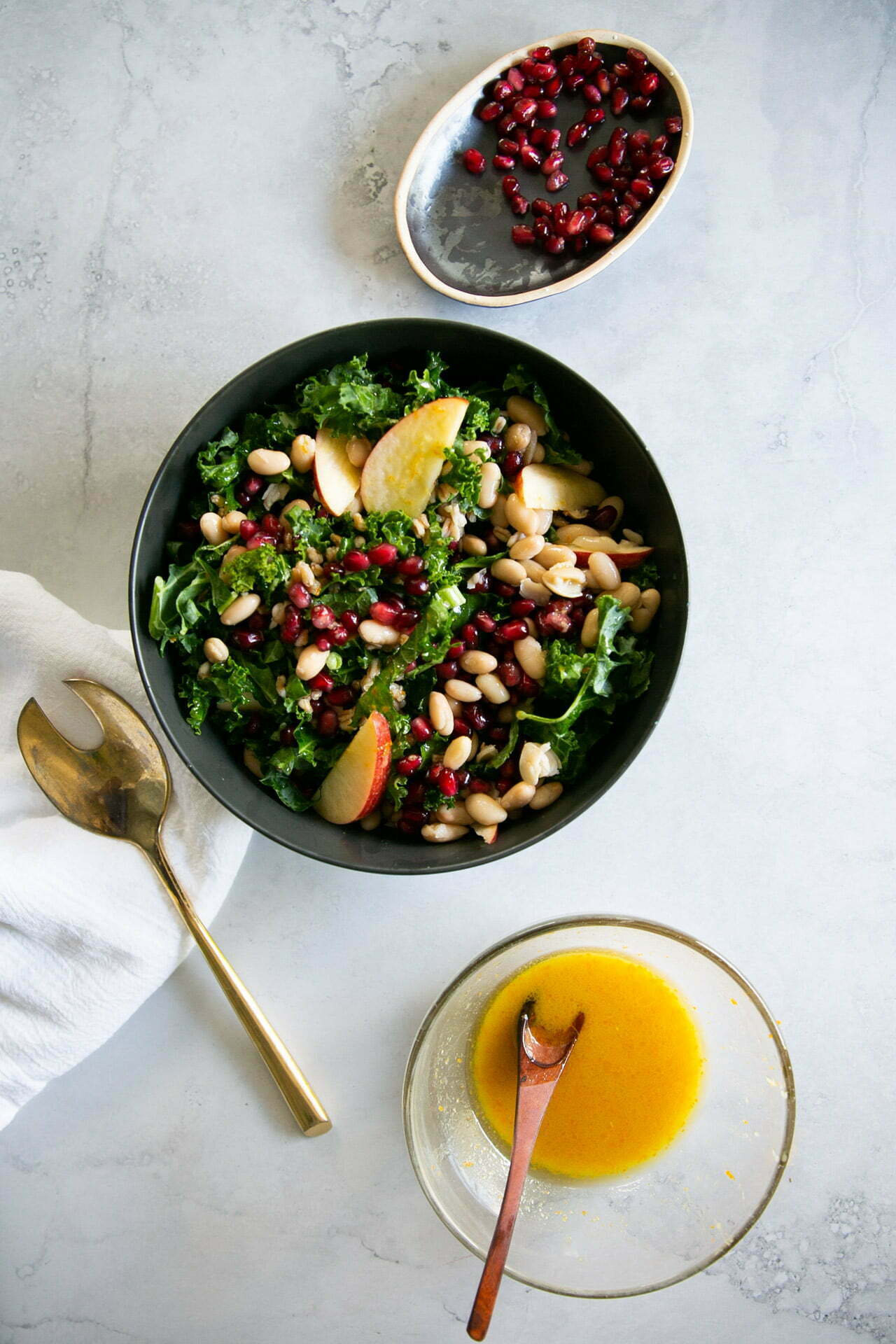 A black bowl filled with a salad of kale and navy beans, apple slices, and pomegranate seeds sits on a light surface. Nearby are a small dish of pomegranate seeds, a bowl of yellow dressing with a wooden spoon, a white napkin, and a gold salad fork.