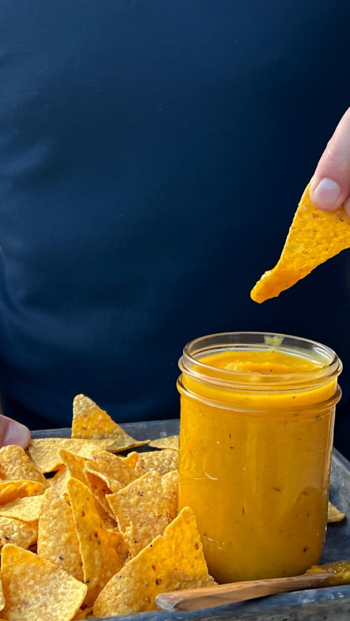 ROASTED HABANERO MANGO SALSA in a mason jar with tortilla chips and a hand dipping a chip into it.