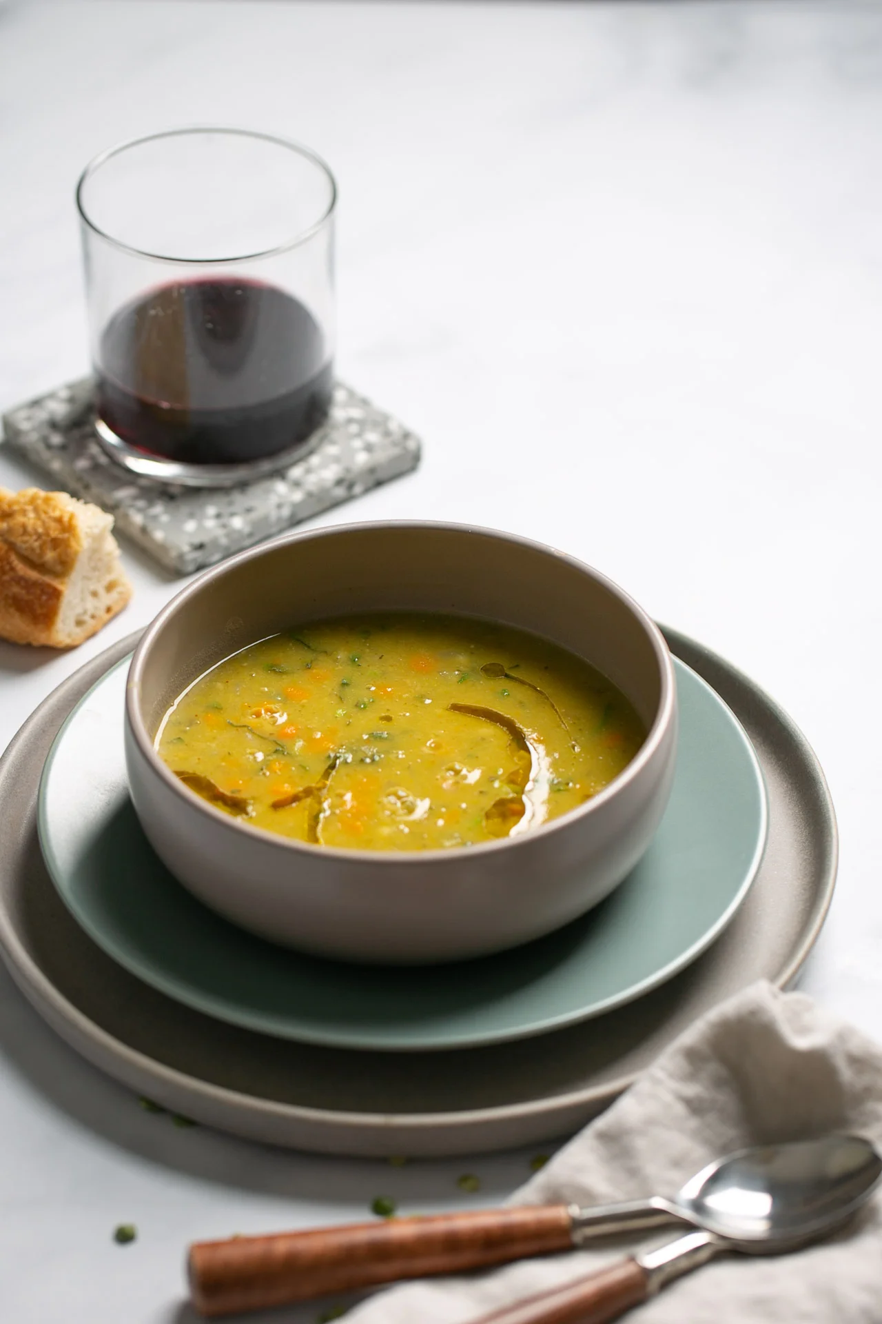 A bowl of yellow split pea soup with herbs sits on stacked plates. Next to it are a glass of red wine on a coaster, a piece of bread, and a folded napkin with a spoon and fork, all arranged on a light-colored table.