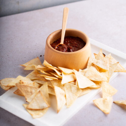 A beige bowl filled with chunky guajillo salsa and a wooden spoon sits on a rectangular white tray. Surrounding the bowl are numerous golden-brown, triangular tortilla chips. The tray rests on a light, speckled surface, while the background is softly textured and neutral-colored.