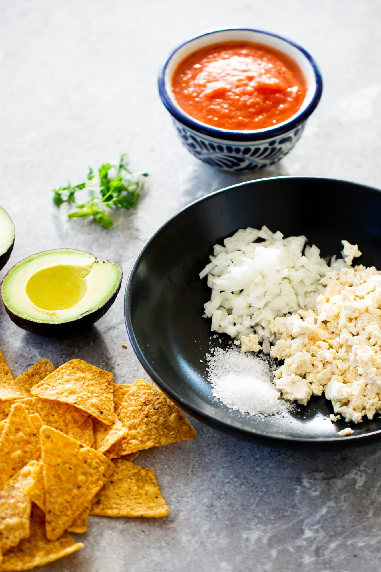A black bowl holds chopped white onion, crumbled white cheese, and a small pile of salt. Nearby are a halved avocado, tortilla chips, fresh cilantro, a small blue bowl of red salsa&mdash;all perfect alongside your favorite overnight oats recipe.
