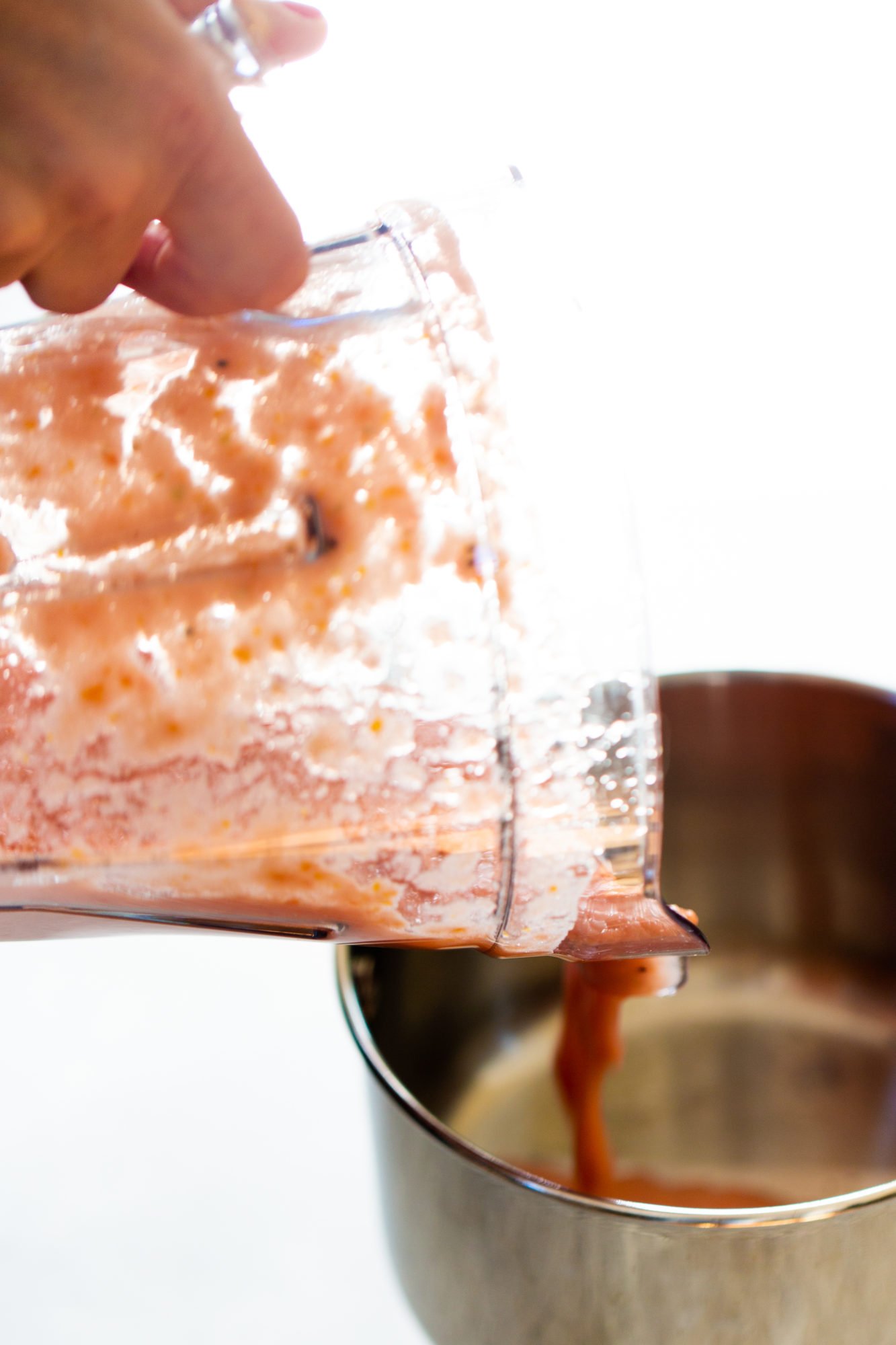 A hand tips a blender jar, pouring thick, reddish-orange puree&mdash;perfect for an overnight oats recipe&mdash;into a stainless steel bowl. The jar is mostly empty but coated with puree, and the brightly lit background remains out of focus.