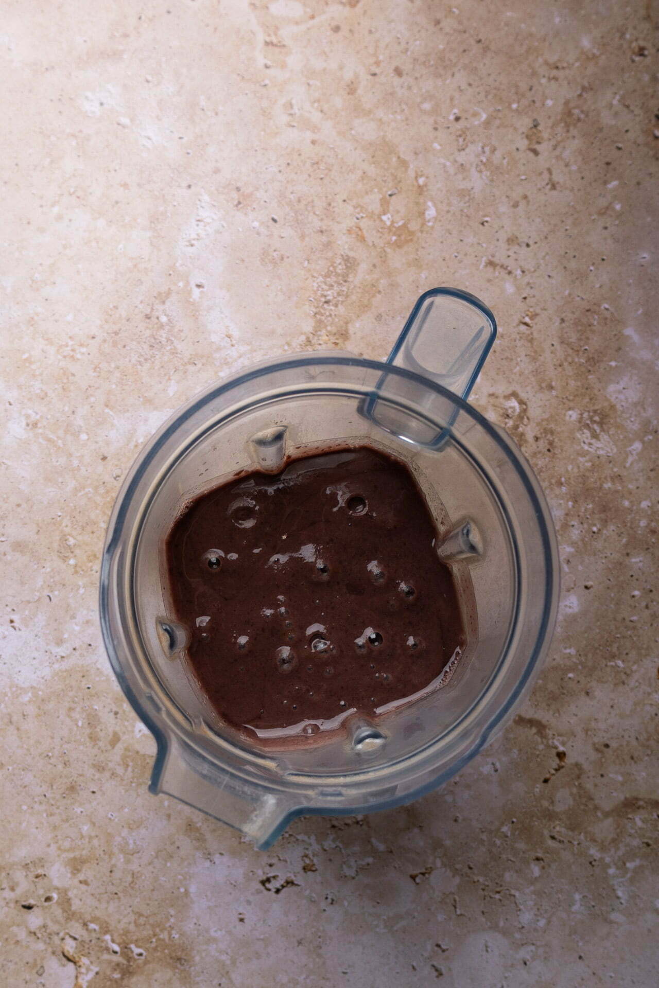 A top view of a blender containing a thick, smooth dark brown chocolate mixture reminiscent of rich Mexican loaded beans. The blender sits on a light brown stone countertop, tiny air bubbles dotting the surface. Its transparent handle is positioned slightly off-center, revealing the luscious blend inside.