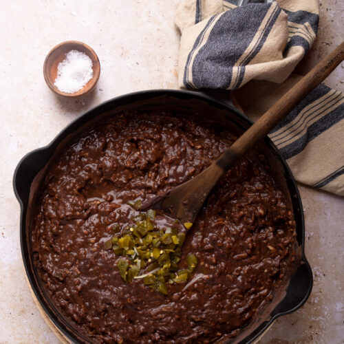A skillet brimming with chili con carne and Mexican loaded beans is topped with chopped green chilies. A wooden spoon rests inside the dish, while a small bowl of salt and a striped kitchen towel lie on the marbled surface, all enveloped in an earthy, warm-toned color scheme.