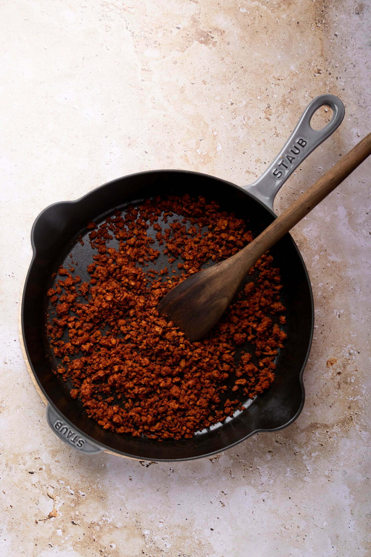 A cast iron skillet with cooked ground meat or a meat substitute sits on a textured stone countertop, ready for Mexican loaded beans. The mixture is browned and crumbly. A wooden spoon rests in the skillet, used for stirring. The skillet has raised logo branding on the handle and is slightly angled.