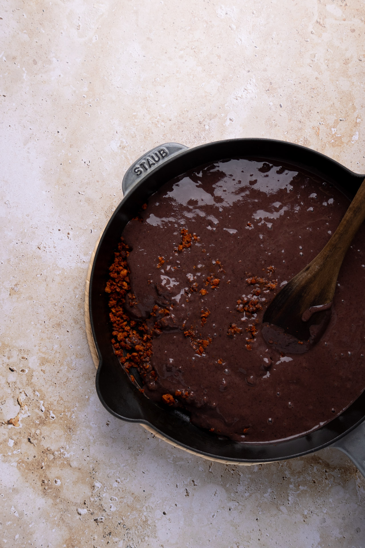 A black skillet filled with rich, dark mole sauce and chunks of chorizo is complemented by a side of frijoles puercos. A wooden spoon sits partially submerged in the mixture, all atop a beige textured surface. The deep color of the mole contrasts beautifully with the light background.