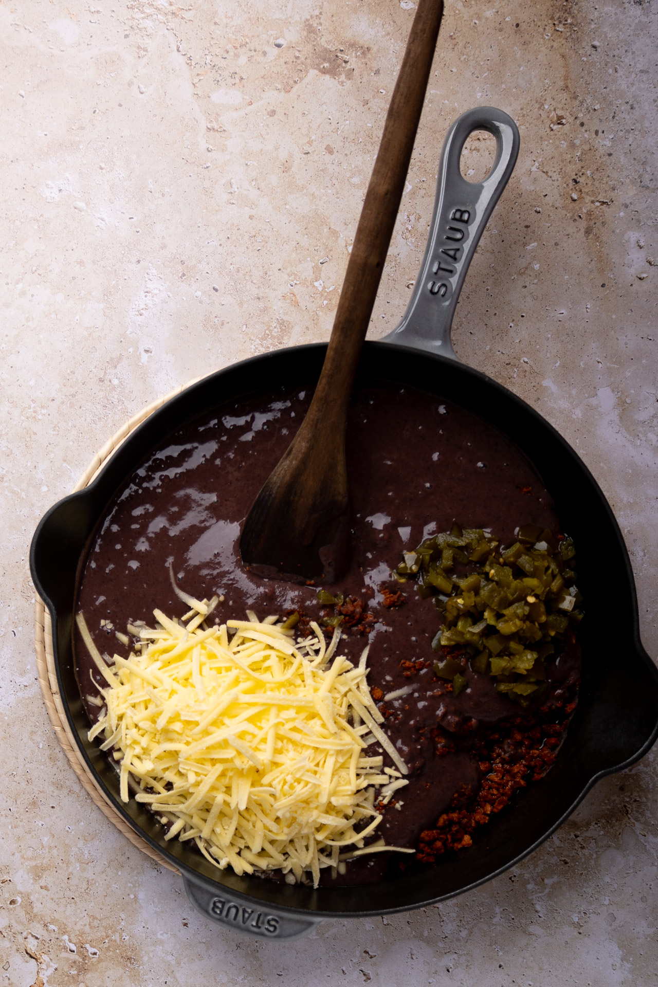 A cast iron skillet showcases a delectable blend of Mexican loaded beans, featuring black bean puree crowned with shredded cheese, chopped green chilies, and red chili flakes. A wooden spoon rests on the edge, atop a textured, light-colored surface.