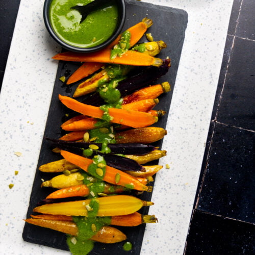 A rectangular slate plate holds an artful array of roasted carrots in vibrant shades of orange, yellow, and purple, elegantly drizzled with green sauce. A small black bowl brimming with extra green sauce accompanies them. The slate rests on a larger white speckled plate atop a dark tiled surface.