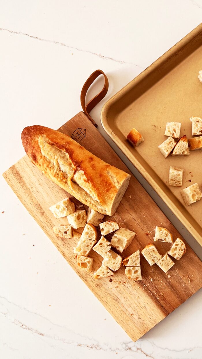 Cutting board with a piece of french bread cut ino cubes and a baking sheet next to it.