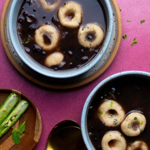 Two bowls of black bean soup with masa harina dumplings rest on a vibrant pink surface. Each bowl brims with rich, dark broth and herbs. A wooden plate displays sliced cucumbers and mint leaves, while a gold spoon nearby adds a touch of elegance to the setting.