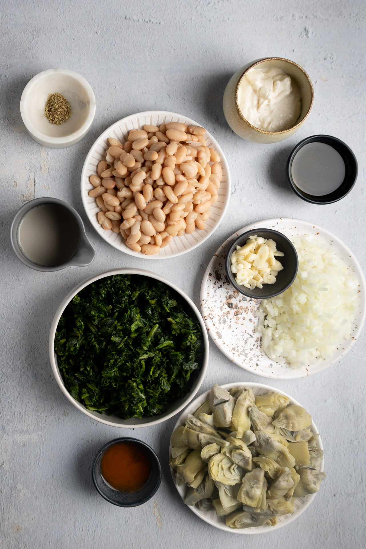 A flat lay of bowls containing vegan artichoke hearts, white beans, chopped onions, minced garlic, chopped spinach, white sauce, broth, olive oil, seasonings, and a small bowl of pepper arranged on a light gray surface.