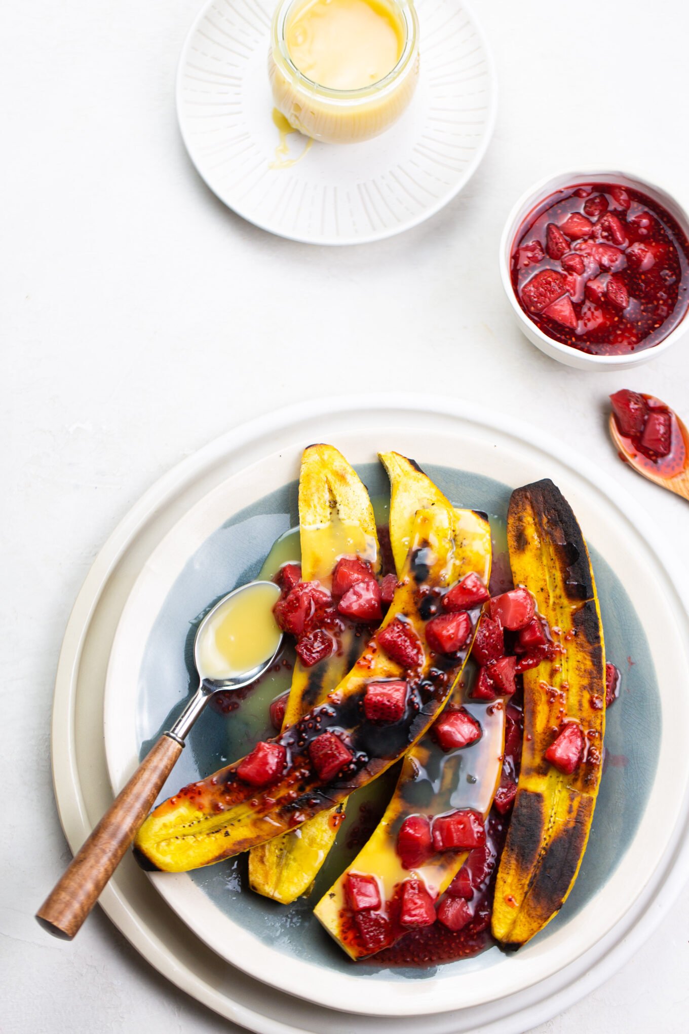 Grilled sweet plantain halves topped with diced strawberries and drizzled with a creamy sauce create a delightful Mexican dessert. A cup of extra strawberries and a spoonful of sauce accompany them. In the background, a small glass with more sauce sits gracefully on a saucer.
