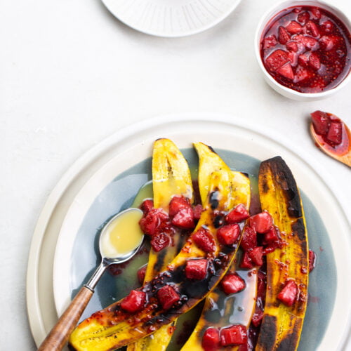 Grilled sweet plantain halves topped with diced strawberries and drizzled with a creamy sauce create a delightful Mexican dessert. A cup of extra strawberries and a spoonful of sauce accompany them. In the background, a small glass with more sauce sits gracefully on a saucer.